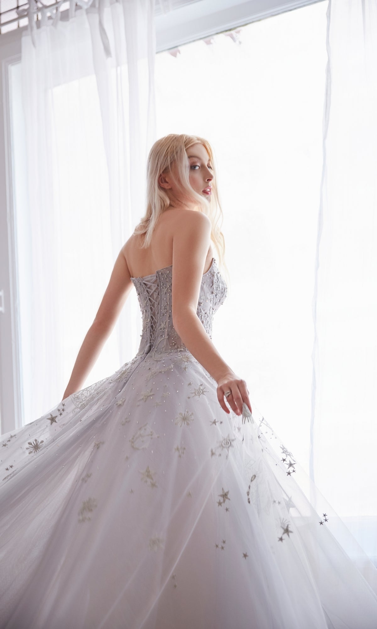 A woman with long blonde hair wears the Constellation-Print Strapless Prom Ball Gown A0890, standing by a large window with sheer curtains and looking back over her shoulder.
